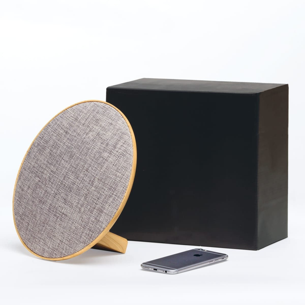 A round, wooden Bluetooth speaker with a grey fabric cover, next to a black box and a smartphone.