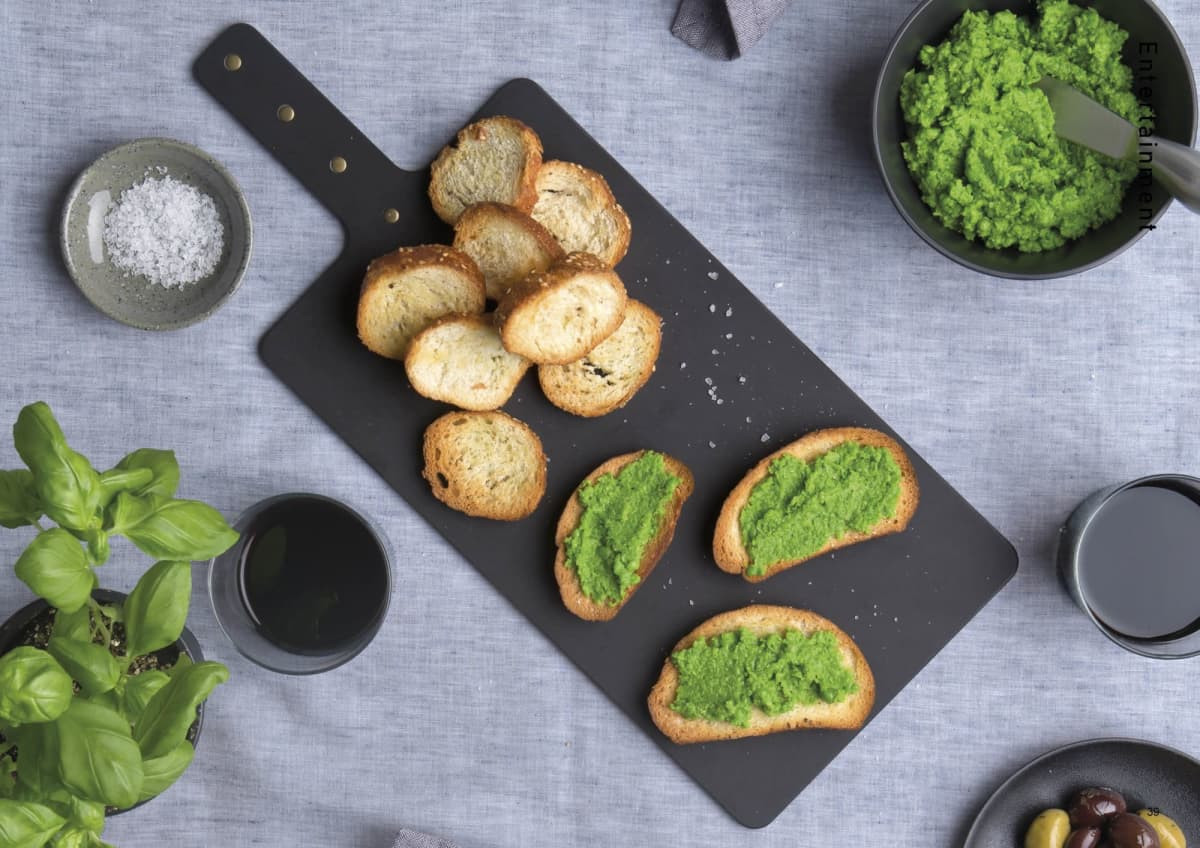 A black Napoli serving board with toasted bread topped with green spread, surrounded by bowls of olives, herbs, and salt.