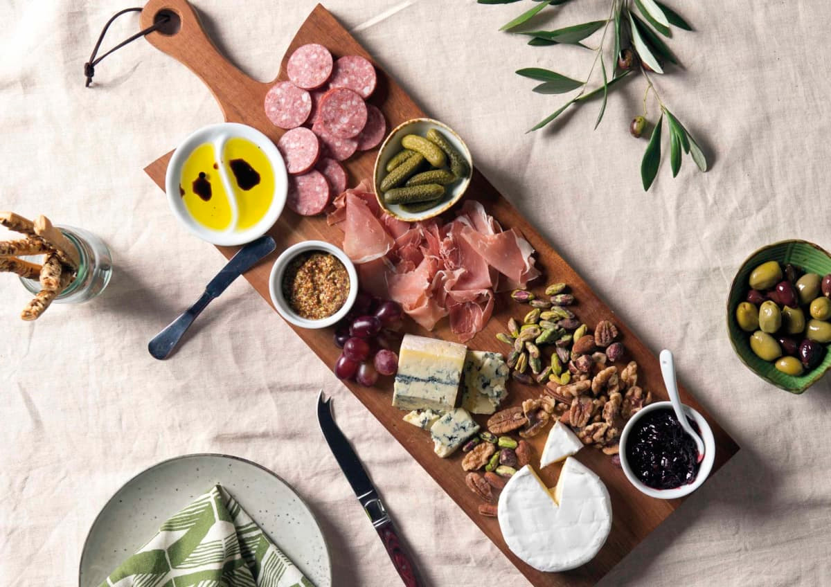 A wooden serving board featuring cured meats, cheeses, olives, pickles, and condiments, with a natural finish.