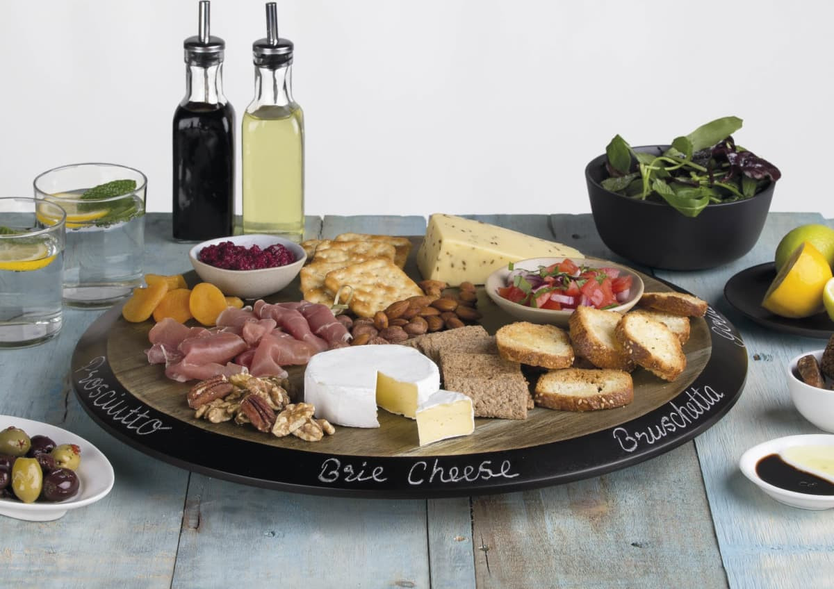 A round wooden serving platter features various cheeses, meats, nuts, and breads, with bowls of olives and salad.