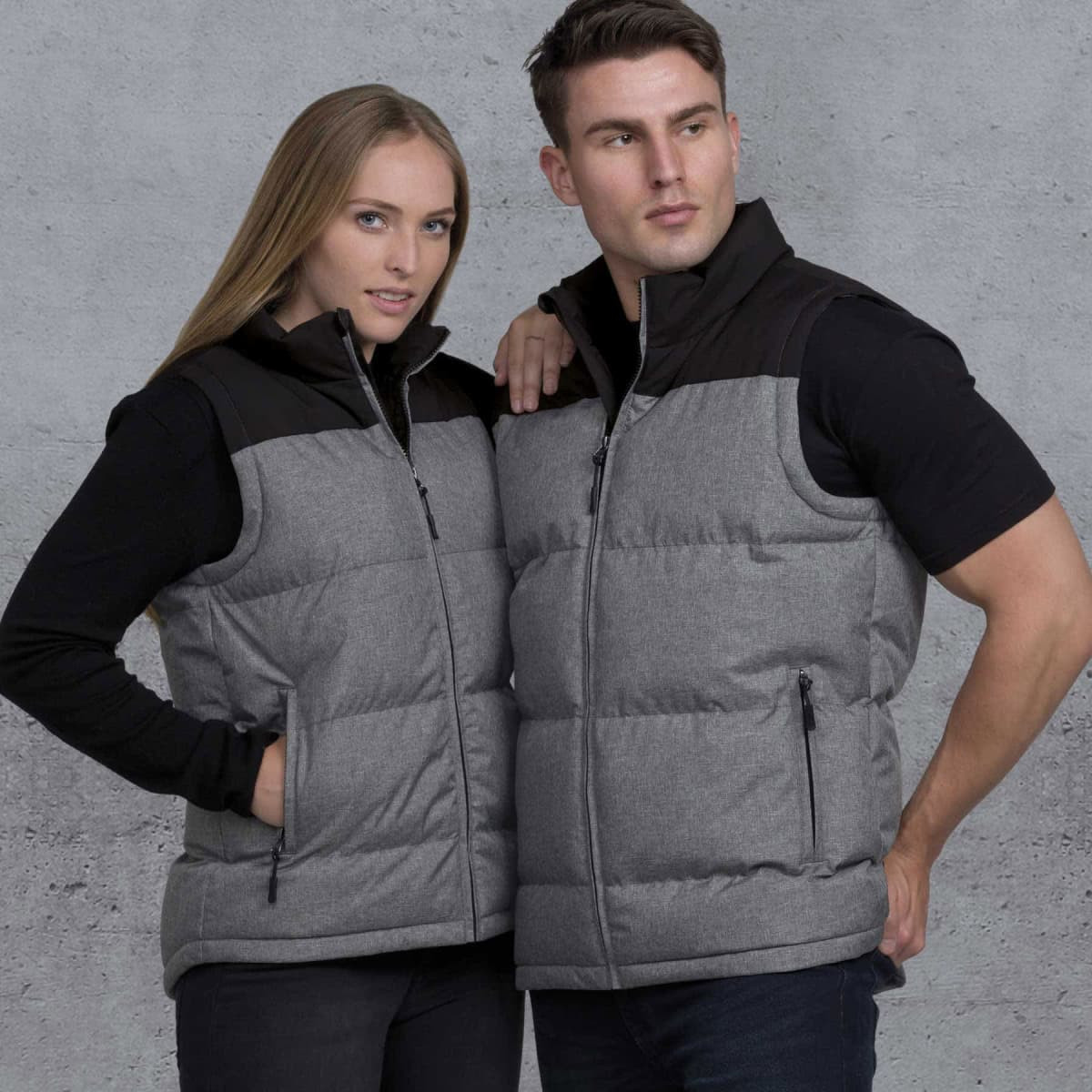 A grey and black puffa vest is worn by a man and woman against a textured background. The vest features a zip and pockets.