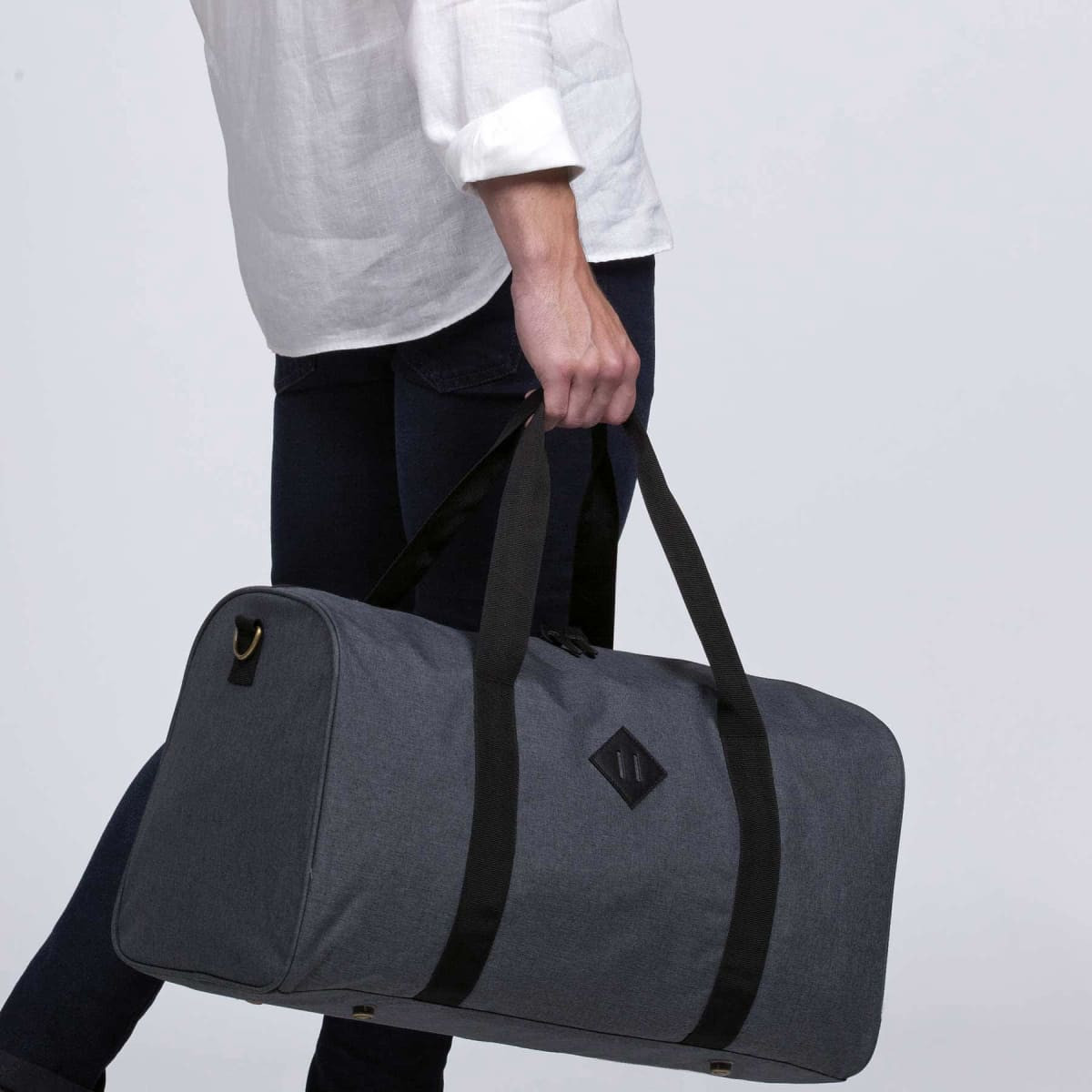 A grey duffle bag with black straps, held by a person wearing a white shirt and dark jeans. It features a logo.