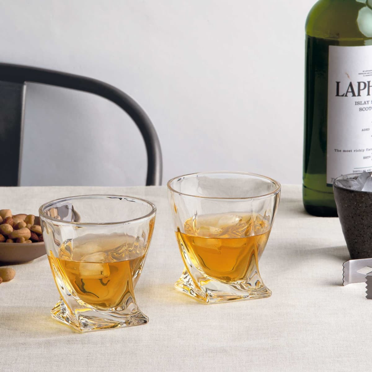 A set of two clear glass whisky tumblers with a unique design, accompanied by a bottle of whisky and snacks.
