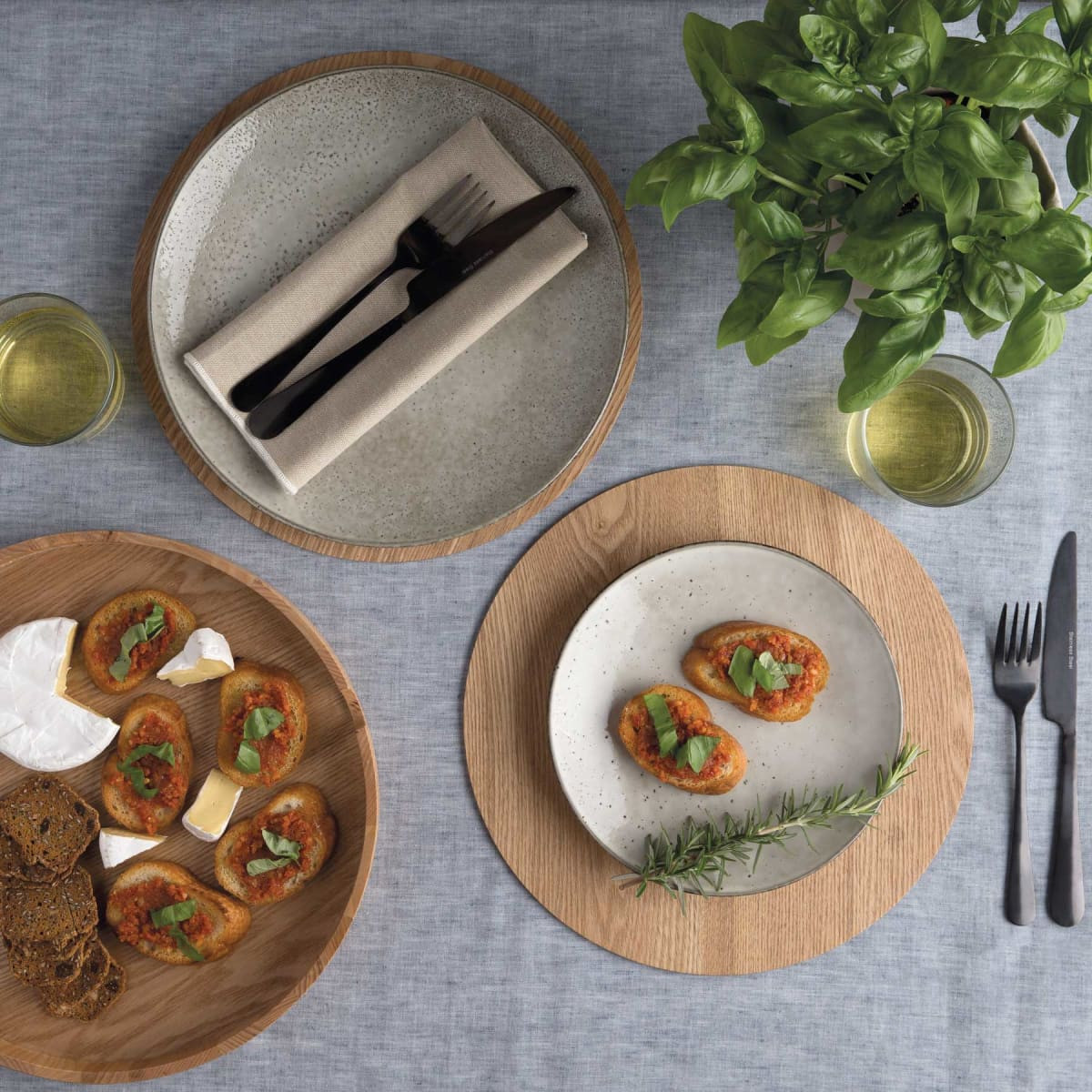 A set of wooden and ceramic placemats featuring food and utensils, complemented by a green plant and glasses of drink.