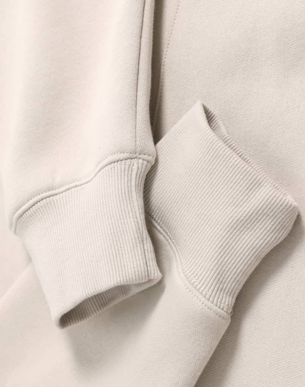 A close-up of a beige oversized hoodie featuring ribbed cuffs and a soft fleece texture. It has a logo.