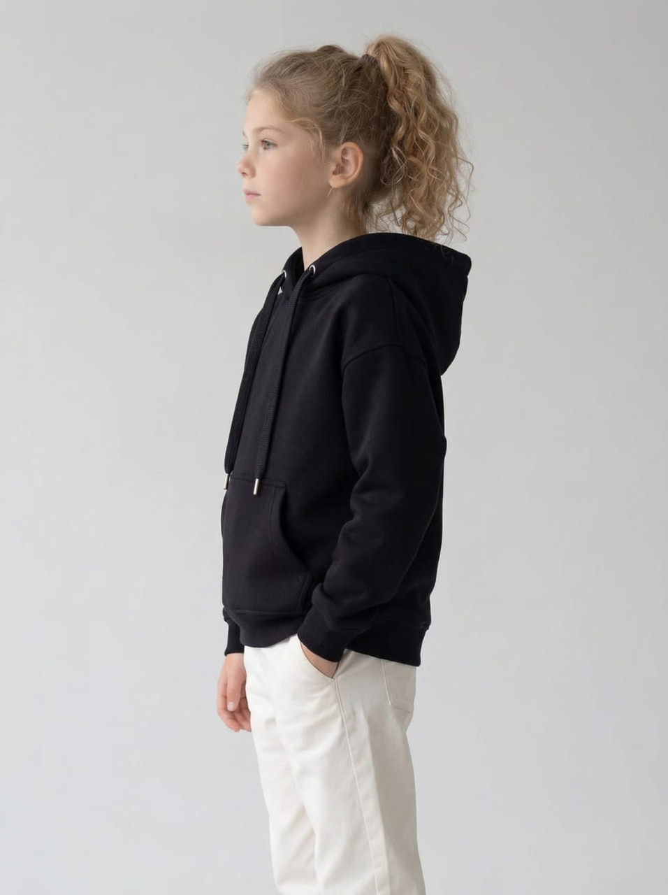 A black hoodie with a front pocket and drawstring, worn by a child standing sideways against a neutral background.