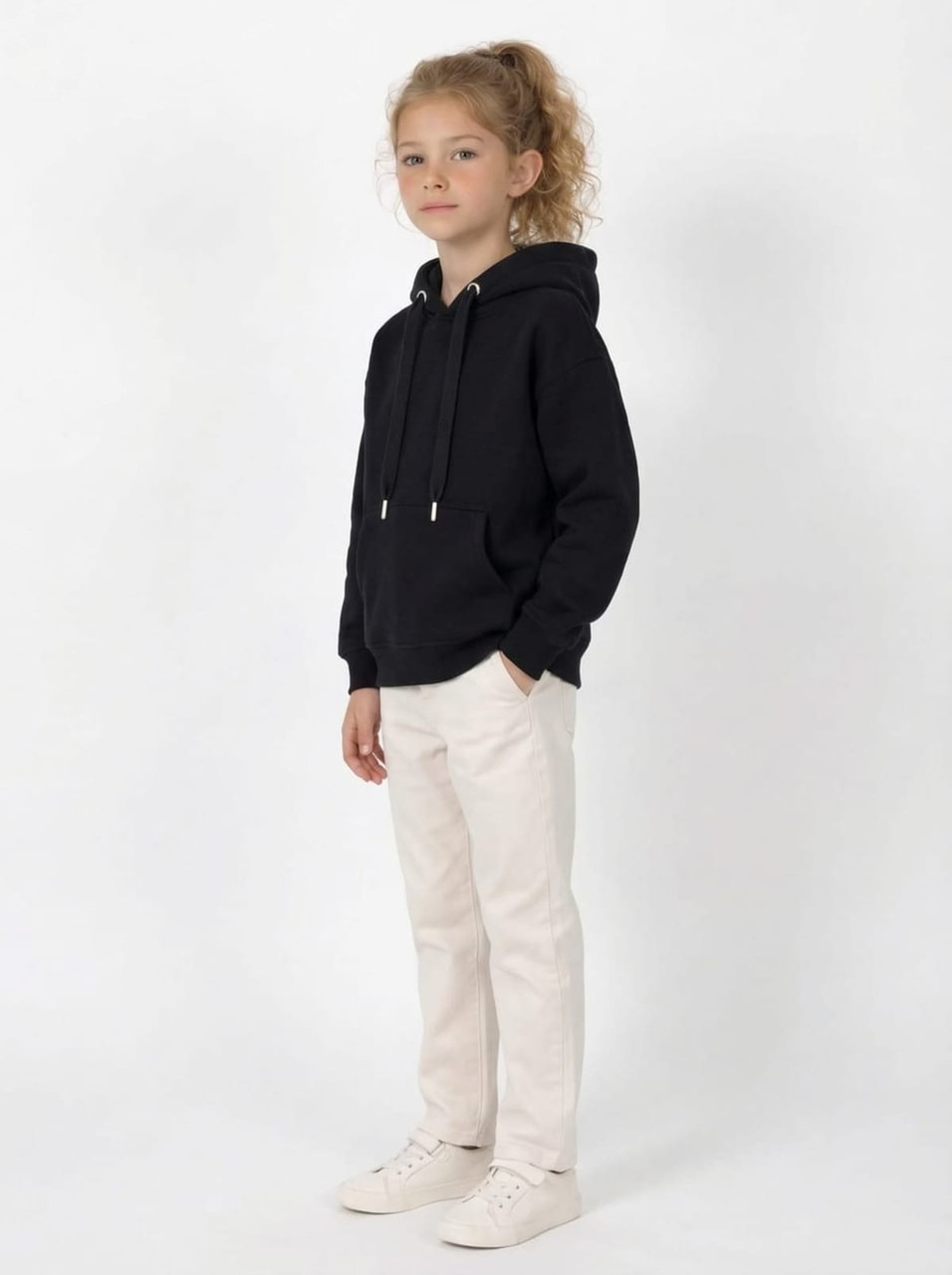 A youth black hoodie with a kangaroo pocket and silver drawstrings, paired with cream-coloured trousers and white shoes.