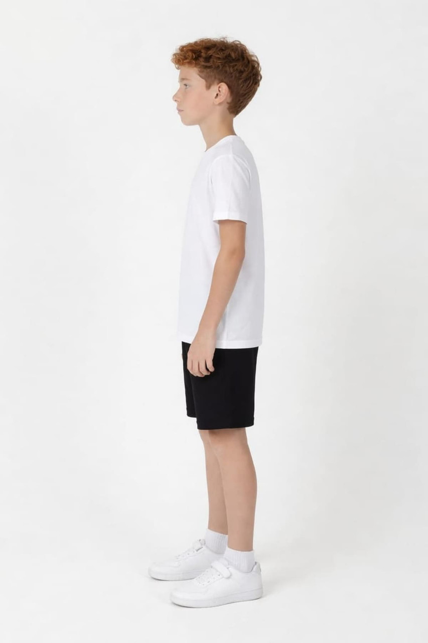 A young boy stands in profile wearing a white t-shirt and black sports shorts with a logo.