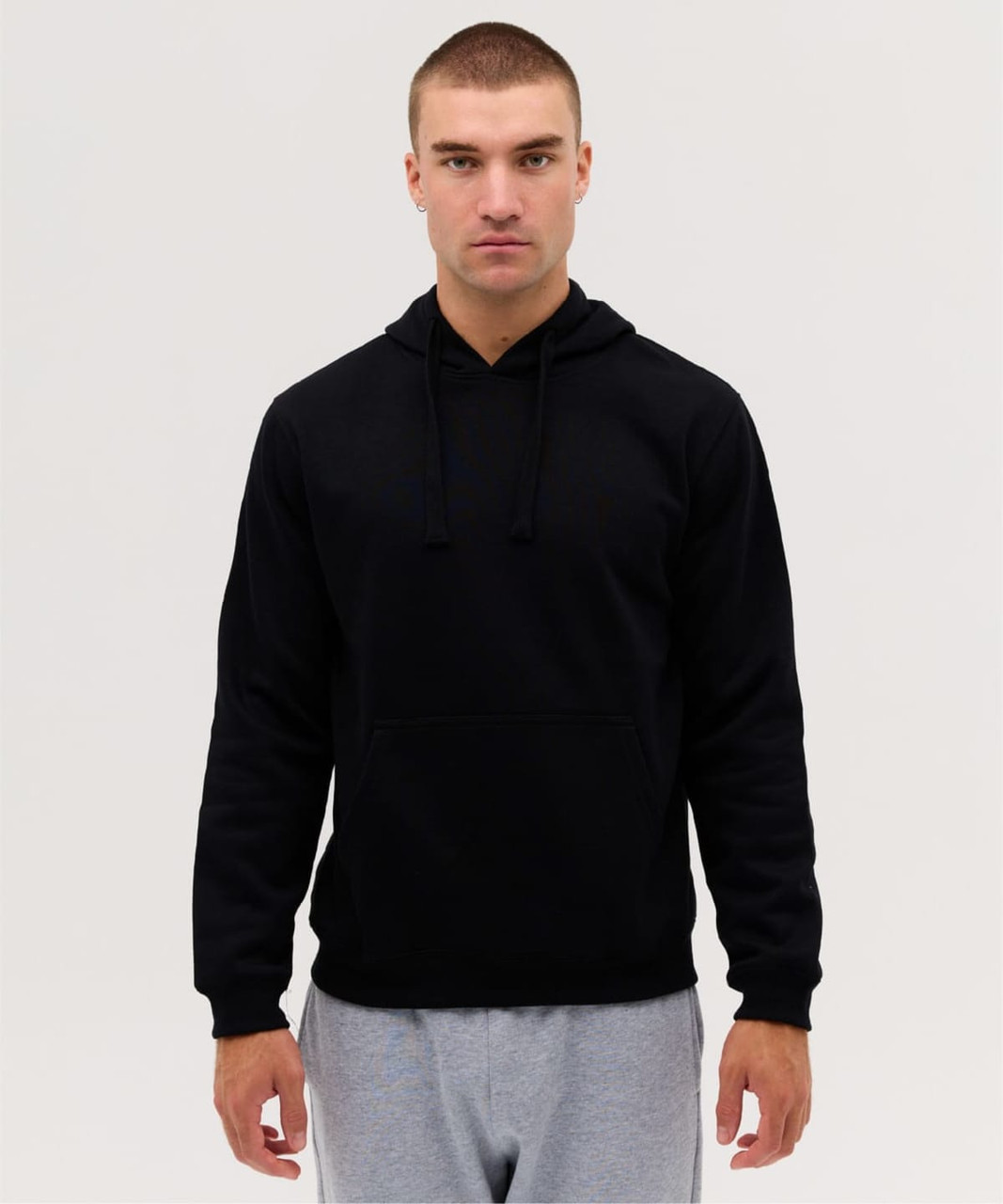 A black hooded jumper with a front pocket and drawstrings, worn by a model. The model is also wearing grey pants.