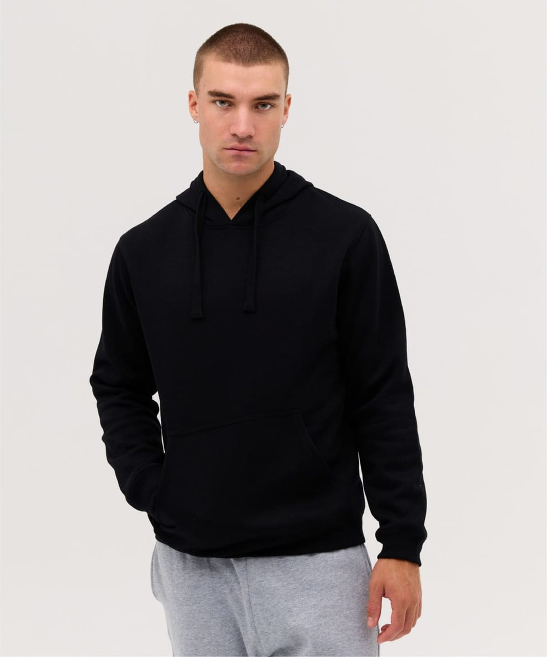 A black hoodie with a front pocket and adjustable drawstring hood, featured on a model wearing grey trousers.