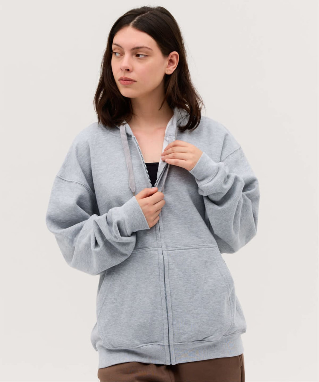 A grey zip-up hoodie with a relaxed fit and a logo. The model has long hair and is posing against a neutral background.
