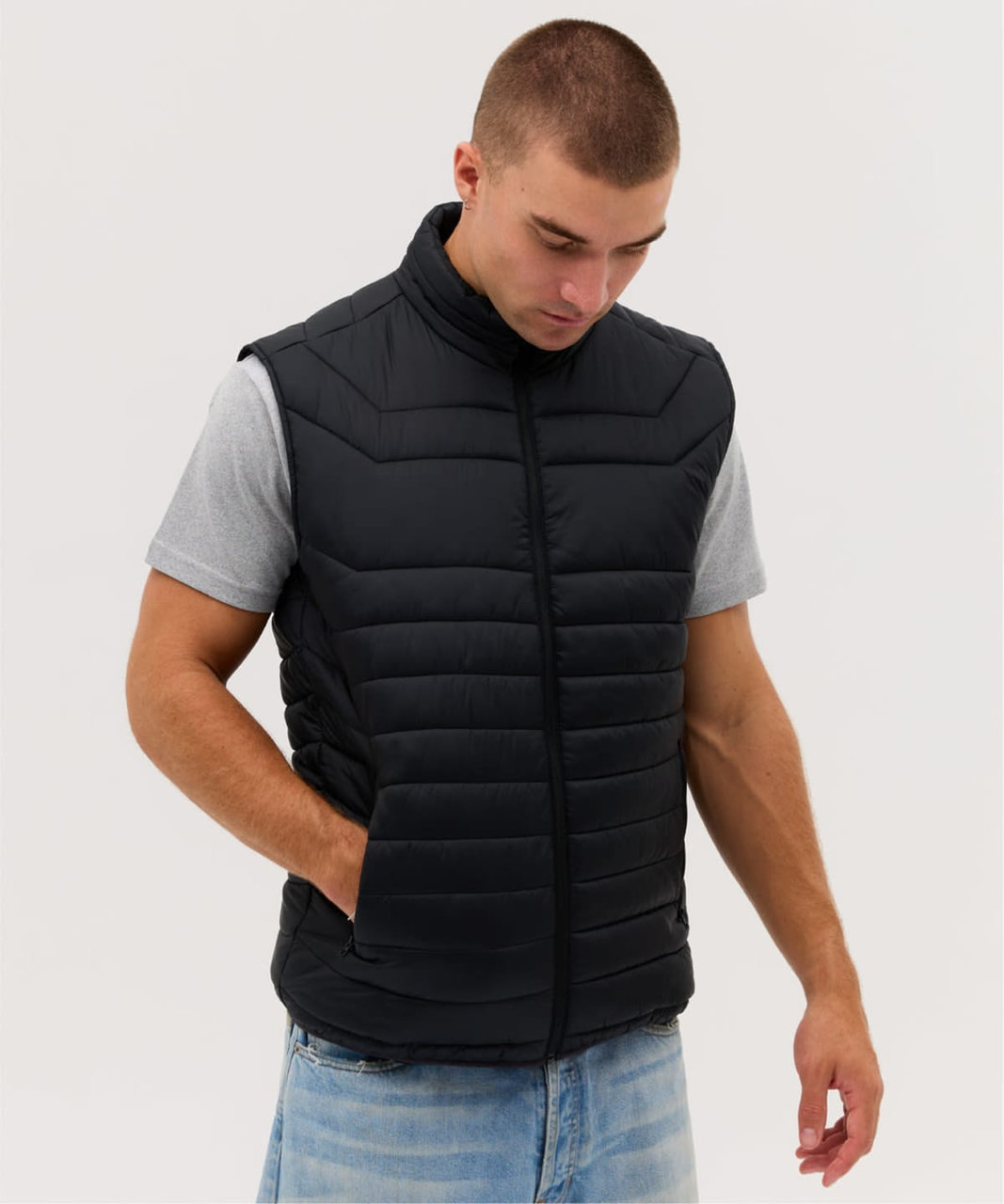 A man wearing a black puffer vest over a grey t-shirt, standing with his hands in the vest's side pockets.