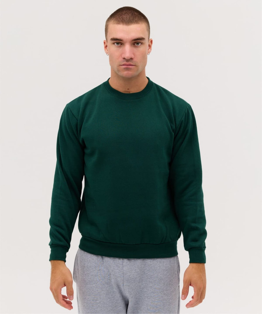 A green crew jumper displayed on a model, featuring long sleeves and a simple design. The model wears grey pants.