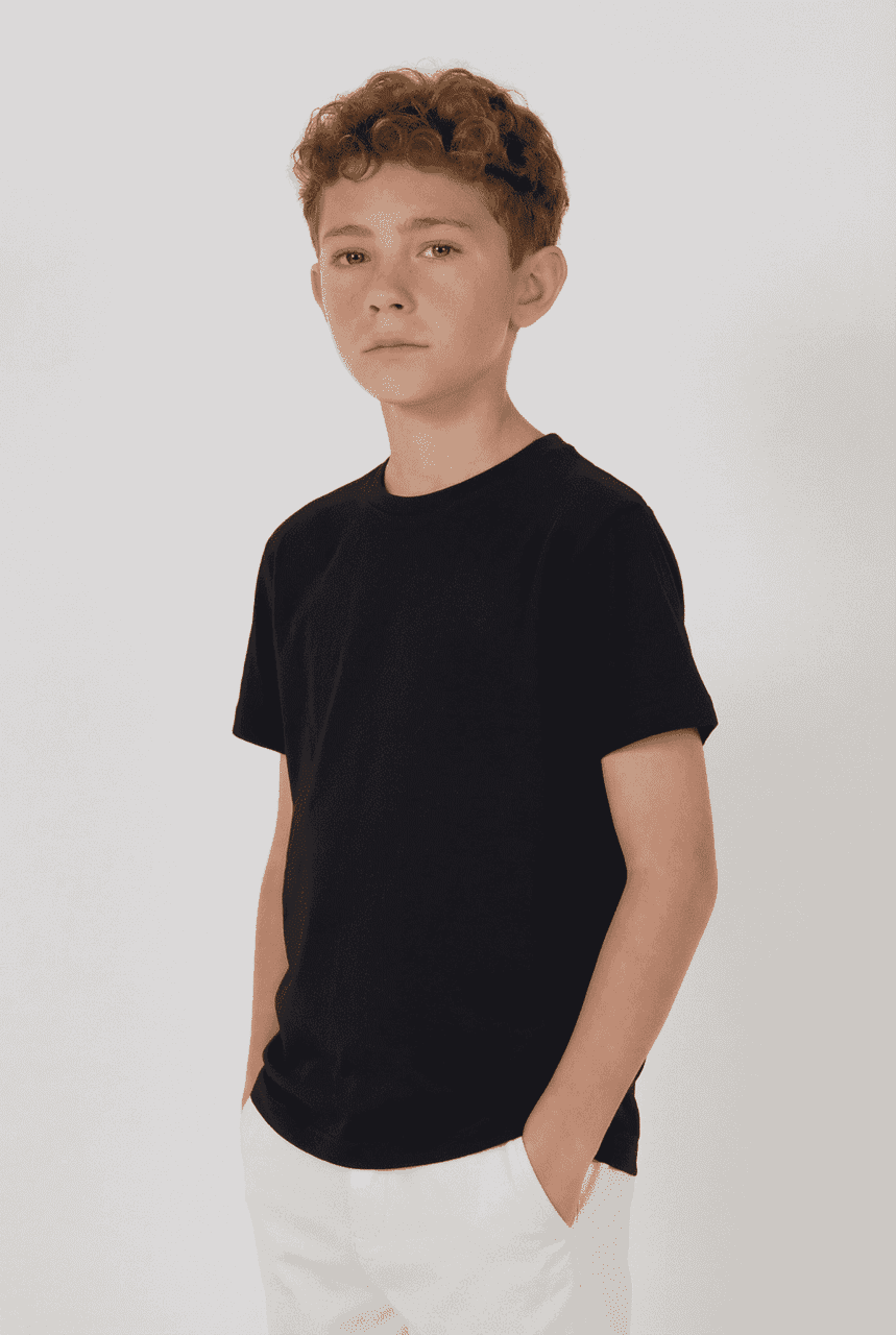 A black youth essential tee displayed on a model, standing with hands in pockets against a neutral background.