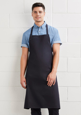 A man wearing a dark navy bib apron over a light blue shirt, standing against a white wall. The apron has a logo.