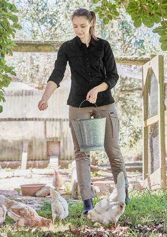 A woman wearing a black long sleeve shirt and beige trousers feeds chickens outdoors, holding a metal bucket.