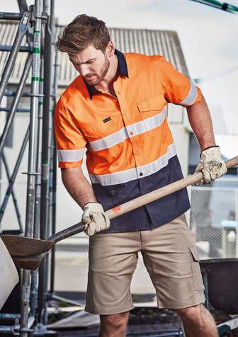A short sleeve work shirt in orange and navy with reflective tape, worn by a man using a shovel, with a construction setting in the background.
