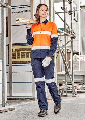A woman in an orange and navy work shirt and matching pants stands on a construction site, holding wooden planks.