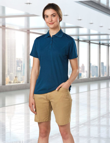 A model wears navy polo and khaki stretch cotton chino shorts in a modern indoor setting. The shorts have a logo.