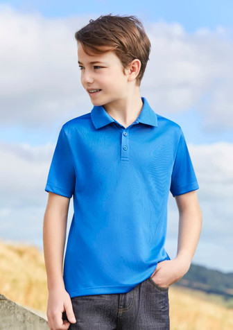 A blue short sleeve polo shirt on a young boy, standing outdoors with a scenic background. The shirt has a logo.