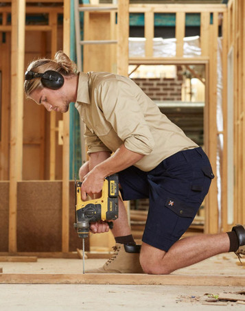 A man in khaki and navy work shorts operating a drill, wearing ear protection, in a construction setting.