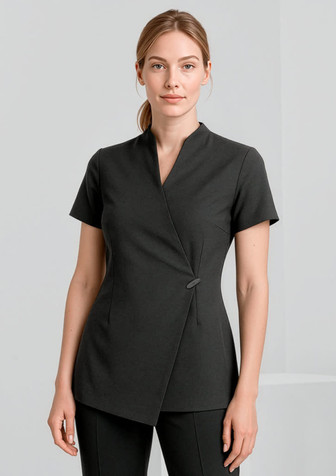 A woman is wearing a black spa tunic with a wrap design and short sleeves. The tunic features a simple button detail.