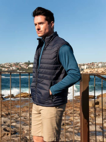 A navy puffer vest worn over a long-sleeve blue top, with a scenic coastal background. The model stands by a railing.