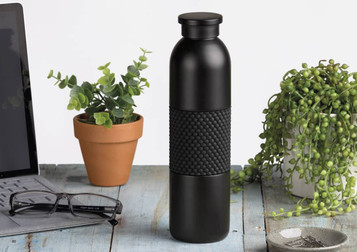 A drink bottle in matte black with a textured black band, placed on a wooden surface alongside plants and office items.