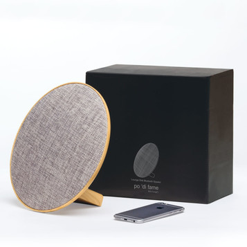 A round grey Bluetooth speaker with a wooden base next to a smartphone, accompanied by a black box packaging.