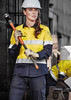 A woman in a yellow and navy long sleeve work shirt, wearing gloves and a hard hat, holding a tool on a construction site.
