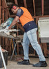 A man wearing an orange and navy short sleeve polo shirt works with a saw in a workshop setting.