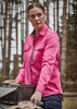 A women's long sleeve button-up shirt in vibrant pink, featuring a logo, set against a forest backdrop.