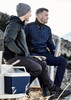 A grey and navy unisex hoodie is worn by two men sitting outdoors, one holding a drink bottle and a cooler nearby.