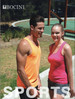Two models wearing singlets, one in orange and the other in pink, standing outdoors with a green area in the background.