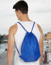 A blue swim backpack with drawstring closure, worn on the back of a person, showing a scenic waterfront.
