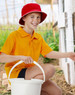 A red bucket hat sits on a boy wearing an orange polo shirt, holding a white bucket in a garden setting.