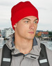 A red beanie worn by a person, paired with a grey jacket, set against a blurred background of a waterfront.