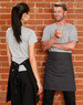 A grey canvas short waist apron in black worn by a person facing another person in casual attire against a brick wall.
