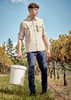 A man in lightweight navy cargo pants and a beige shirt holds a bucket in a vineyard setting, with trees in the background.