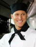 A chef wearing a black scarf and black cap, smiling in a kitchen setting. The scarf complements his white chef's uniform.