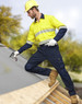 A man wearing a yellow and navy utility shirt and navy cargo pants, standing on a roof, holding a tool.