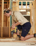 A man in khaki and navy work shorts operating a drill, wearing ear protection, in a construction setting.