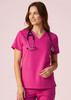 A women's pink V-neck scrub top is displayed, featuring short sleeves and a classic fit, with a stethoscope around the neck.