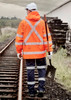 An orange waterproof jacket with reflective stripes, worn by a person holding a shovel, walking along railway tracks.