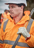 A man wearing an orange waterproof jacket with reflective strips, gloves, and a safety helmet, holding a tool.