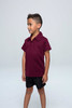 A maroon children's polo shirt with a collar, displayed on a young boy wearing black shorts. The shirt has a logo.