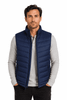 A man models a navy blue puffer vest over a light grey long-sleeve shirt, featuring a branded logo.