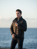 A black puffer jacket for men, featuring a zip-up front and a logo, worn by a model against a coastal backdrop.