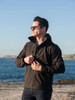 A man wearing a black soft shell jacket stands by the water, adjusting his jacket with sunglasses on.