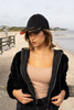 A black cap with a hi-vis orange peak worn by a woman on a beach, in a casual outfit and standing near a boardwalk.